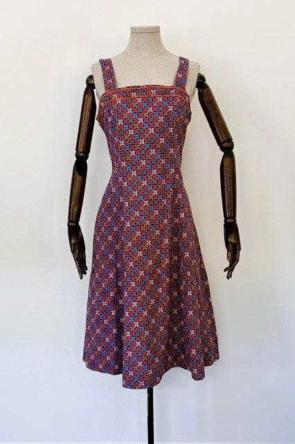 A sleeveless fit-and-flare dress with a geometric pattern in red, blue, and white, displayed on a mannequin with dark wooden arms against a plain white background.
