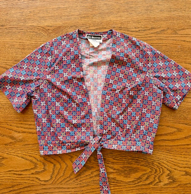 A short-sleeve cropped wrap top with a tie-front hem, featuring a dense geometric floral pattern in red, blue, pink, and white. The brand name 'THE MARSH' is visible on the inner neck label.