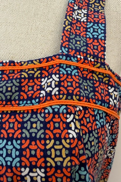 Close-up of a fabric tote bag with a navy blue background and a colorful geometric pattern in orange, light blue, and white. The bag features bright orange piping along the seams and a matching strap, shown against a neutral beige textured fabric backdrop.
