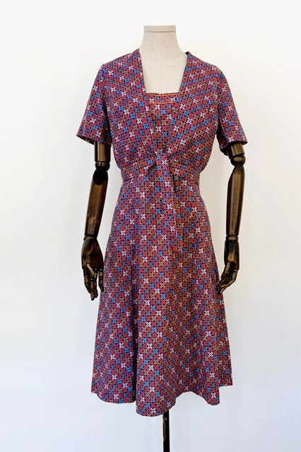 A short-sleeve knee-length dress with a geometric pattern in red, blue, and white, displayed on a mannequin with wooden arms against a plain white background.